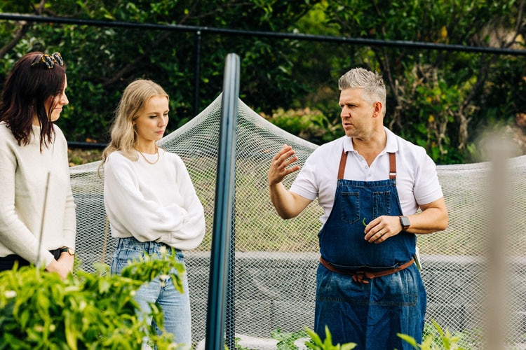 massimo mele wearing a blue apron talking to two women about his vegetable garden